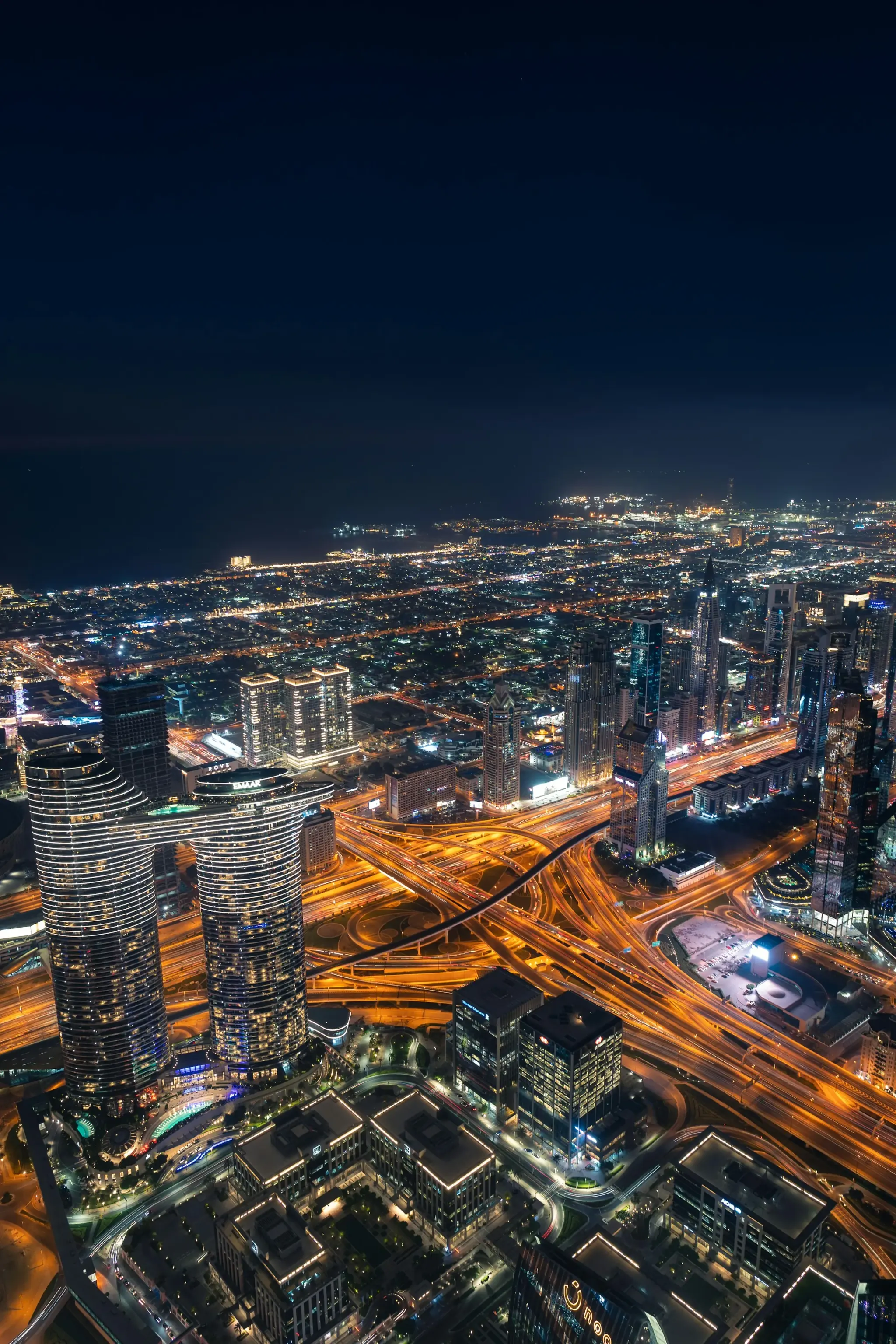Dubai at night from high above Downtown: the interchange traced in light below, towers and the lit grid of the city running away to the coast.