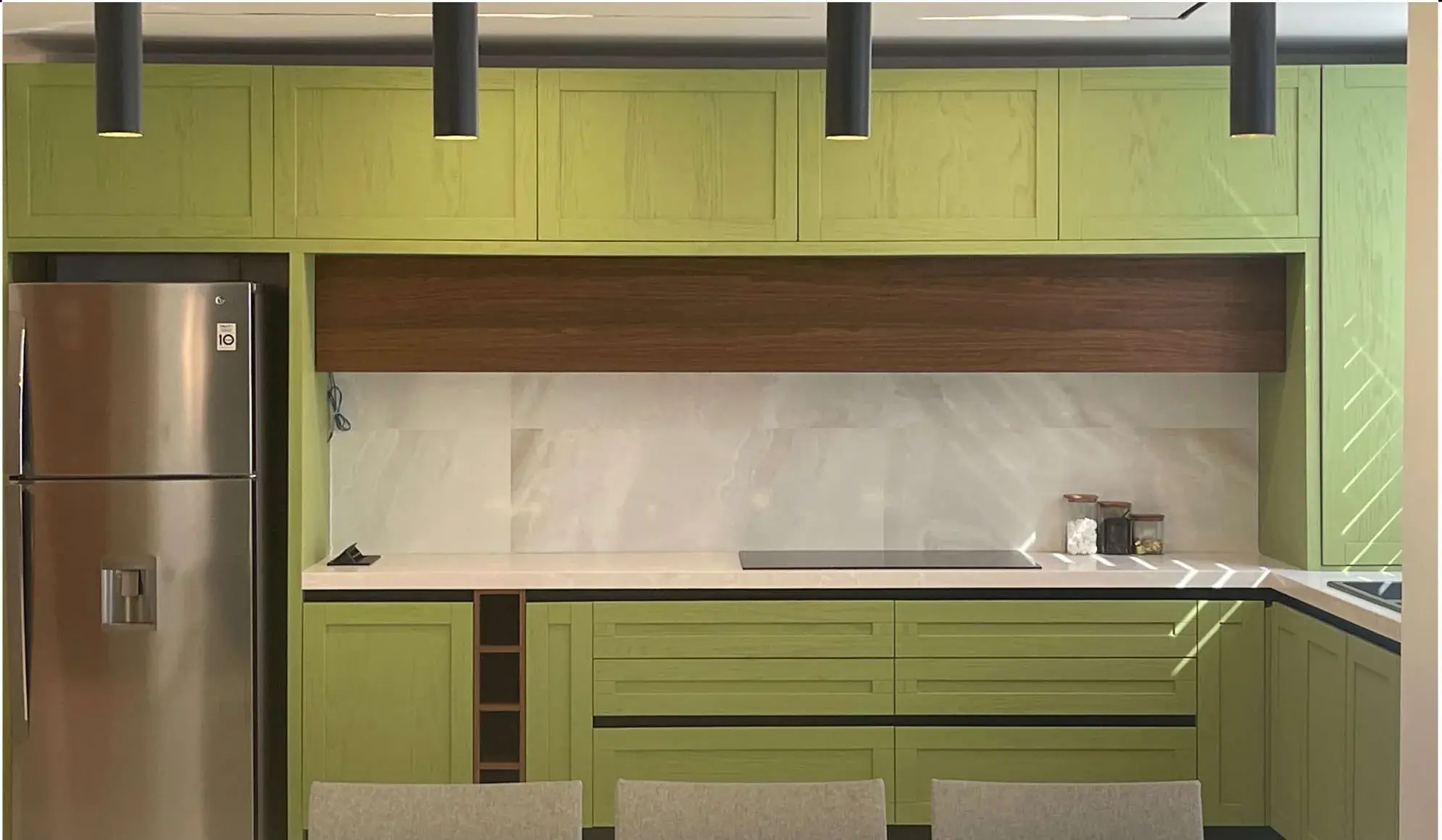 Green lacquered kitchen photographed straight on, with a walnut soffit niche above the worktop and cylindrical pendants over a breakfast counter.