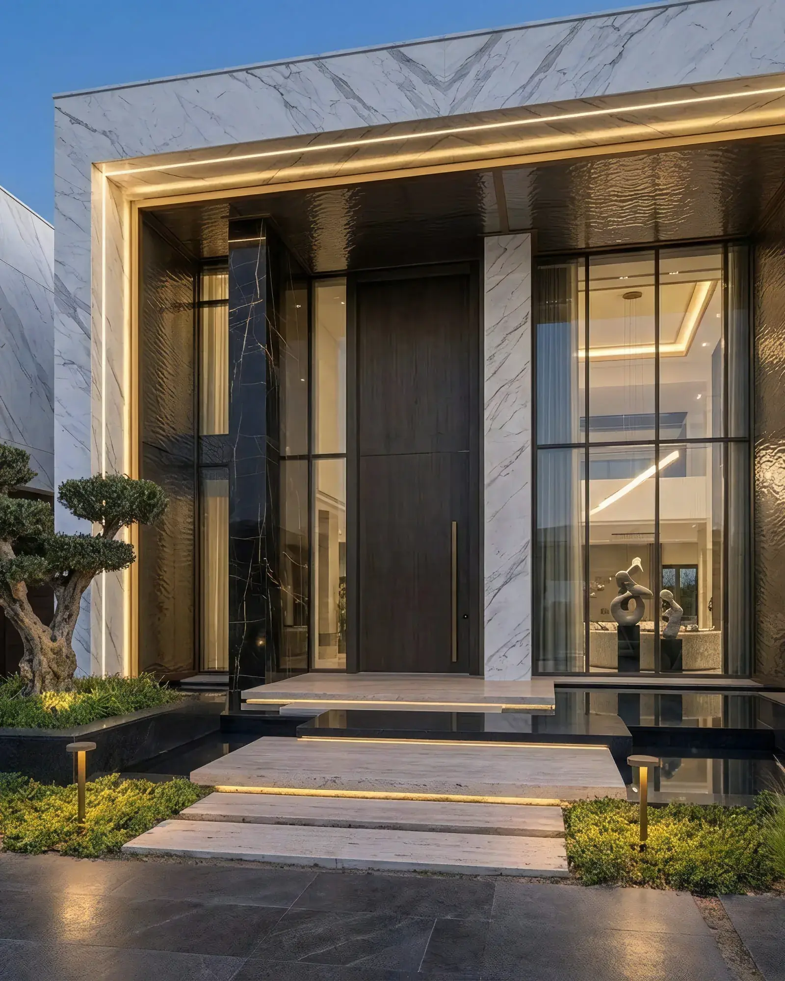 The marble portal seen square on: a recessed reveal lit along its full perimeter, a book-matched black marble blade to one side of the pivoting timber door, and the glazed foyer visible straight through to the interior.