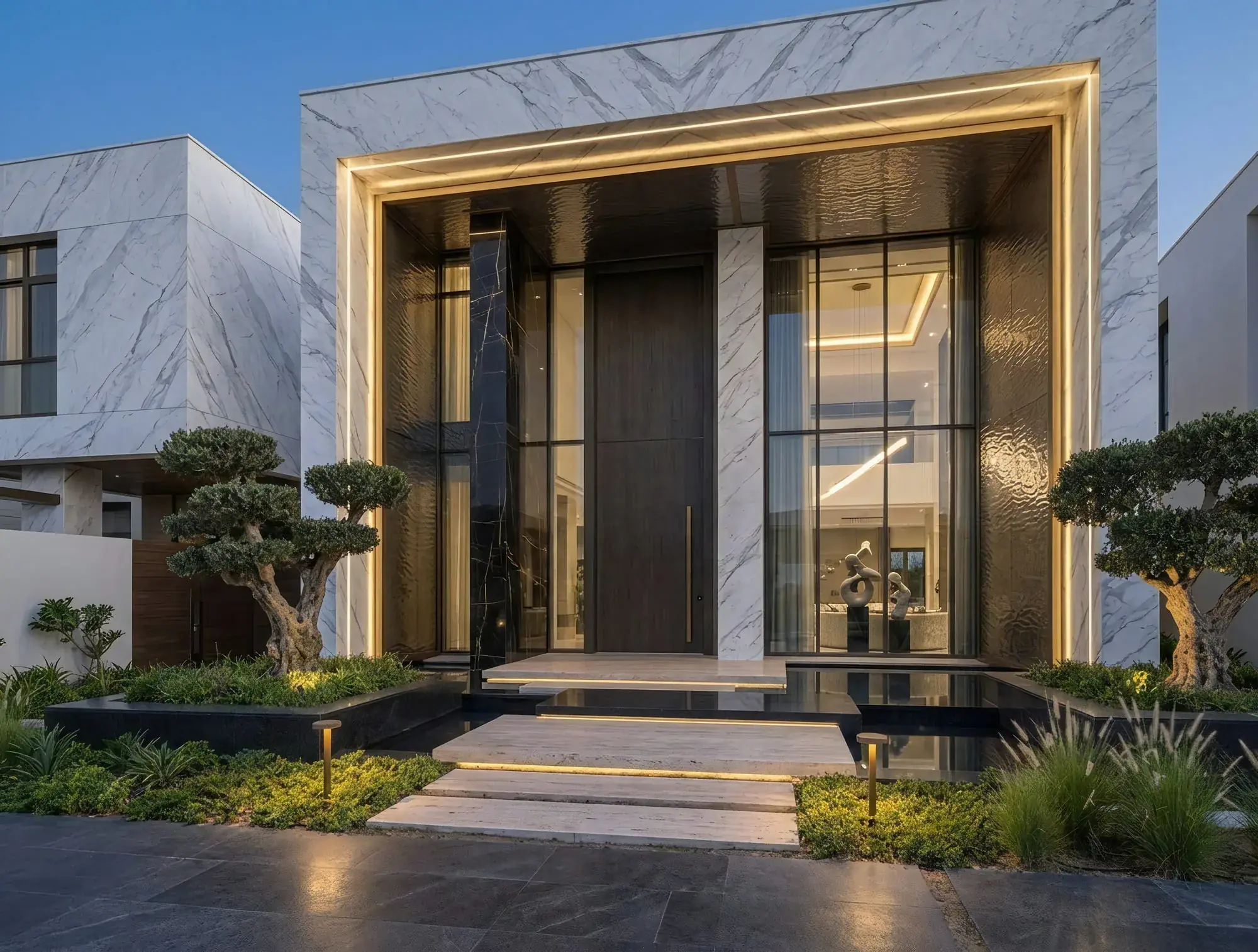 The entrance to Villa Luce at dusk: a double-height marble portal framing a pivoting timber door, with cloud-pruned olives and a black reflecting pool at the threshold.