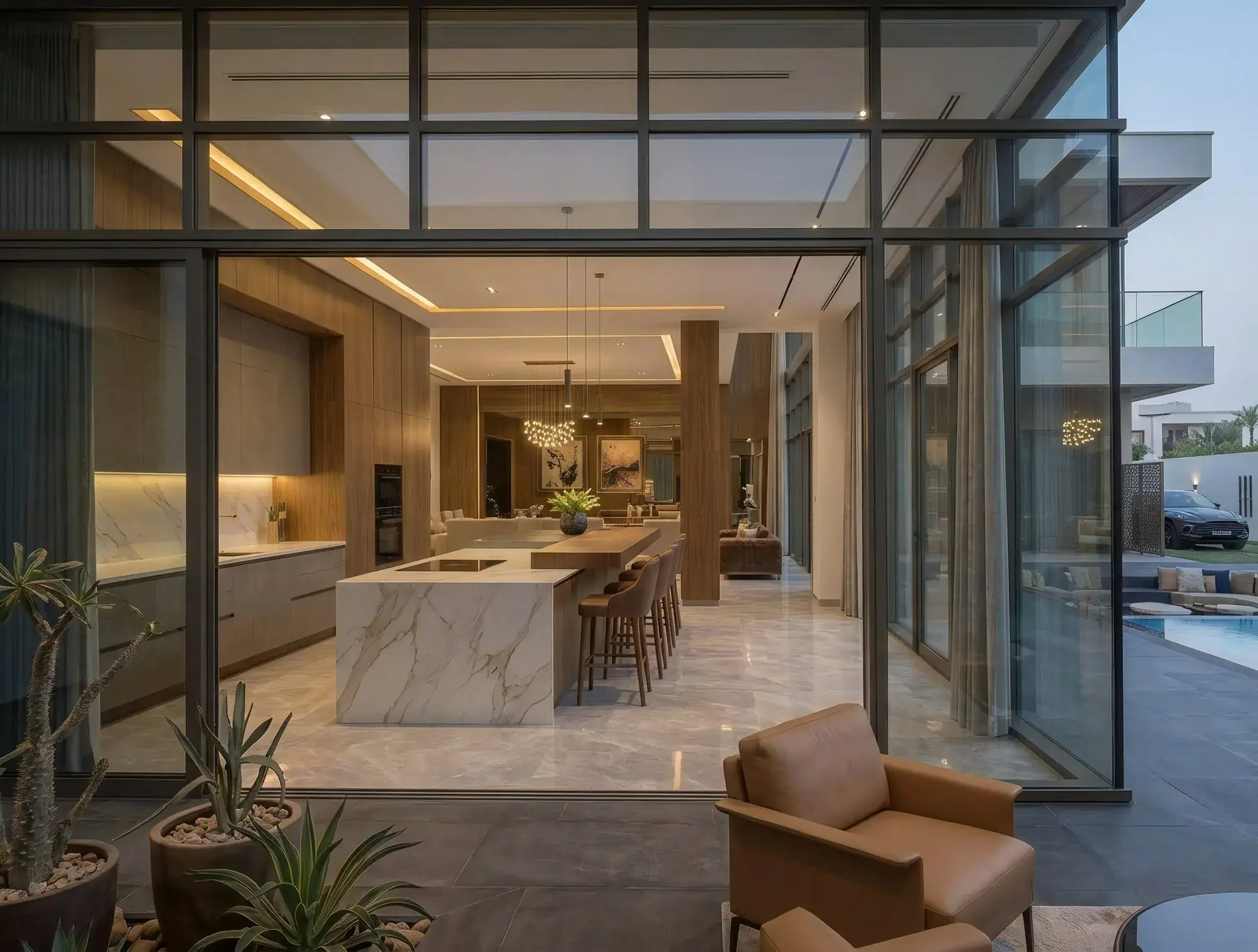 The same kitchen and living space seen from the terrace through full-height sliding glass, with an armchair and planted pots in the foreground and the pool to one side.